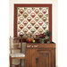 A decorative quilt featuring basket patterns hangs above a wooden cabinet and chair with a plaid seat.