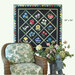 A floral quilt hangs above a wicker chair adorned with a green-patterned cushion and potted flowers.