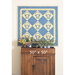 A blue and yellow quilt featuring basket motifs hangs above a wooden cabinet with greenery.