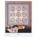 A decorative quilt featuring floral patterns arranged in a grid, displayed above a table with fruit and pottery.