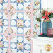 A colorful quilt featuring floral and patterned squares hangs beside a vase of pink roses on a table..