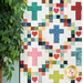 A colorful quilt featuring a pattern of crosses and hearts, displayed beside a leafy plant..