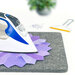 An iron rests on a gray pressing mat with colorful quilt pieces, with scattered pins and a wool mat cleaning tool in the background.