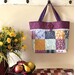 A colorful patchwork tote bag hangs on a wall above a basket of fruit and a bouquet of yellow flowers.