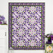 The completed Fresh Cut Roses Quilt colored in white, purple, and green prints from the Plum Petals fabric collection, hung on a white paneled wall and bordered by coordinating florals and decor.