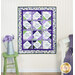 The completed Forever Sisters Charm Quilt colored in vibrant purple, green, and cream fabrics from the Georgina collection, hung on a cream paneled wall and staged with coordinating flowers and furniture.