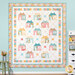 The completed Maisons de Patchwork Quilt colored in bright yellow, pink, and blue fabrics, hung on a blue paneled wall and bordered by coordinating summer decor.
