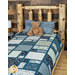 A patchwork quilt in blue tones is staged on a wooden bed with a plush teddy bear. 