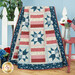 A patriotic quilt with star patterns in red, blue, and cream draped over a white picket fence next to flowers and coordinating decor. 