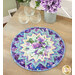 The Point of View Garden Delight table topper featuring a floral star pattern, placed on a wooden table beside three glass cups and purple flowers.