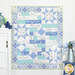 The completed Comfort of Psalms Quilt colored in blue, aqua, white, green, and purple fabrics from the Daydreams collection, staged on a white paneled wall with coordinating decor and furniture.