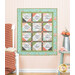 The completed Forever Sisters Charm Quilt colored in vibrant aqua, green, charcoal, and pink fabrics from the Abloom collection, hung on a cream paneled wall and staged with coordinating flowers and furniture.