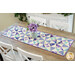 A colorful quilted table runner with geometric patterns, featuring a vase of purple flowers, on a wooden table.