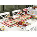 The completed table runner made with log cabin blocks of red, pink, cream, and green floral fabrics from the Regal Rose collection, laying in the center of a white dining table with white chairs and four place settings at each seat with matching cloth napkins.