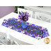 The completed Easy Striped Table Runner colored in vibrant purple floral fabrics from the Pansy Perfection collection, staged on a clean white table with coordinating flowers.