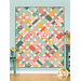 The completed Picnic Quilt colored in vibrant, peach, gold, aqua, and teal fabrics from the Tango collection by Moda Fabrics. The project is hung on a paneled aqua wall and staged with coordinating flowers and furniture.