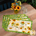 The four completed Self-Binding Placemats colored in the cheerful Sunflower Farm collection, fanned out on a rustic wooden table top and staged with coordinating decor and flowers.