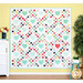 The completed Summer Love Quilt, colored in the bright yellows, reds, and aquas of the Summertime collection, hung on a yellow-paneled wall and bordered by coordinating summer florals and greenery.