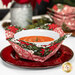 The completed bowl cozy colored in red and green fabrics from the Berry & Pine collection by Moda Fabrics, staged on a red plate and holding a white bowl with a warm pie inside. The other bowl cozies and coordinating decor can be seen in the background.