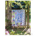 A colorful quilt hangs on a wooden fence gate, surrounded by greenery and sunlight.