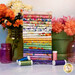 A stack of the fabrics included in the Fleur de Rose FQ Bundle, with jars and a pot and flowers and thread.