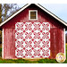 The completed Wish Upon A Star Quilt colored in red and white floral fabrics from the Red Delicious collection, hung from the front of a rustic red barn.