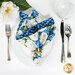 A top down shot of a complete table setting featuring silverware, a glass, and flowers surrounding a white plate with a cloth napkin on the plate, showing off the white floral print of the reversible napkin.