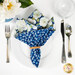 A top down shot of a complete table setting featuring silverware, a glass, and flowers surrounding a white plate with a cloth napkin on the plate, showing off the blue floral print of the reversible napkin.