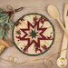 The completed round cream Folded Star Hot Pad, staged on a wooden table with coordinating seasonal and kitchen décor.
