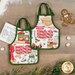 A top down shot of the apron hot pads on a rustic wooden table, staged with treats, holiday greenery, and a spray of flour,