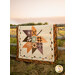 The completed Pressed Leaves quilt colored in autumn fabrics from the The Awe of Autumn collection, hung over a wooden fencepost in front of a field at sunset for a rustic look.