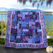 The completed Easy as ABC quilt in purple, blue, and lavender batik fabrics from the Wisteria Blooms collection by Bali Batiks, artfully draped over a railing with a beautiful nature scene in the background. 