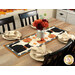The completed Scaredy Cat Table Runner, colored in warm orange, cream, gray, and black fabrics from the Midnight Moon collection, staged on a wooden table with four black chairs and complementary table settings. A galvanized metal tin canister holds coordinating flowers in the center of the runner.