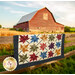 The Fluttering Leaves quilt, colored in colorful fabrics from the Flossie's Flowers collection by Henry Glass and hung over a wooden farm fence. The sun sets on a red barn in the background.