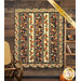 The completed Pumpkin Lane Quilt colored in rich orange, brown, blue, tan, and red fabrics from the Wheatland collection by Moda Fabrics, hung on a rustic wooden wall and staged with coordinating decor between a wooden ladder and vintage rocking chair. A yellow banner in the upper right corner reads 