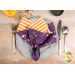 A top down shot of the napkin with the purple side faced out, staged on a gray table setting beside tossed Halloween decor.