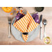 A top down shot of the napkin with the orange side faced out, staged on a gray table setting beside tossed Halloween decor.