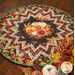 The completed Point of View Folded Star Table Topper colored in vibrant autumn-themed fabrics from the Gather With A Grateful Heart collection by Timeless Treasures Fabrics; pumpkins and coordinating decor peek into frame from the lower right corner.