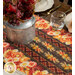 A close up on the middle of the runner, showing details on the vibrant autumn themed fabrics. Coordinating florals in a tin sit in the center and pair with complementary table settings.