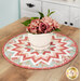 The completed Folded Star Table Topper, colored in pink, blue, and white fabrics from the Muse collection, staged on a wooden table with a pot of flowers in the center of the topper.