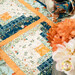 A super close up on one of the log cabin blocks, demonstrating fabric colors and print details as well as top quilting effects. Coordinating flowers and napkins peek into frame from the right side of the frame.