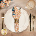 A top down shot of a table setting with a plate, cutlery, a wine glass, and a cloth napkin with the cream fabric with an orange plaid print facing out.