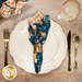 A top down shot of a table setting with a plate, cutlery, a wine glass, and a cloth napkin with the blue floral and pumpkin print facing out.