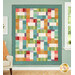 The completed Jelly Sticks Quilt colored in cheery teal, red, gold, pink, and red calico prints from the Gentler Times collection, hung on a blue wall next to a window and armchair.