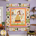 The completed Ghoul Friends panel quilt colored in spooky cream, black, orange, green, and purple fabrics, hung on a purple wall and surrounded by coordinating Halloween décor as a black cat sleeps on a rocking chair next to the quilt.