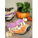 A completed bowl cozy colored in black, orange, cream, and purple, staged on a wooden table with a stack of other bowl cozies and coordinating Halloween décor.