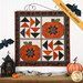The completed Pieceful Patchwork - October wall hanging, a combination of patchwork pumpkin and flying geese blocks colored in cream, orange, and black Halloween fabrics. The project is hung on a white paneled wall and staged with coordinating decor. A banner at the top reads, 