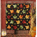 The completed Fall Foliage quilt, colored in contrasting red, orange, green, yellow, and black batik fabrics from the Forest Flora collection, staged on a rustic red barn with pumpkins and cornstalks. 