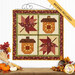 The completed Pieceful Patchwork - November wall hanging, a square project with two patchwork acorns and leaves in autumn themed fabrics, hung on a white paneled wall and staged with coordinating fall decor like corn, pumpkins, and leaves.