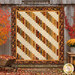 The completed 28th Floor quilt, colored in bold neutral tones like cream, brown, red, and gold, hung on a gray paneled fence, framed by vibrant fall trees and garden decor.